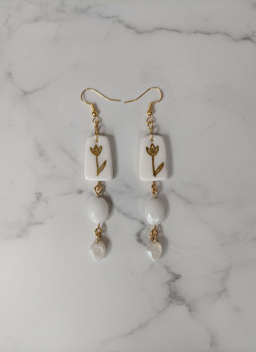 Boucles d'oreilles retro or et blanches avec motif floral peint à la main sur argile blanche, perles en résine et apprêts en acier inoxydable. Design vintage chic de 9 cm pour un style artisanal élégant.