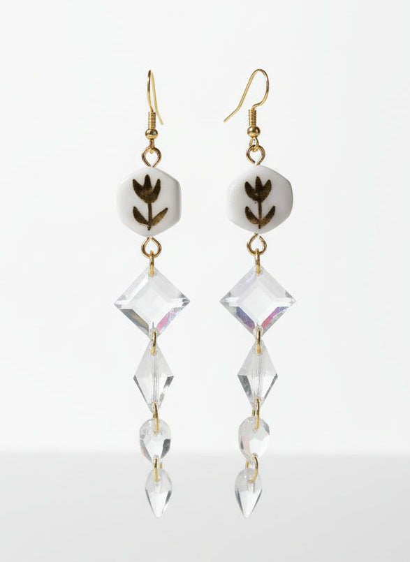 Boucles d'oreilles florales or et blanc avec perles hexagonales blanches décorées de motifs floraux noirs et cristaux transparents suspendus. Bijou fait main chic en acier inoxydable doré, idéal pour une soirée élégante ou un look vintage raffiné.